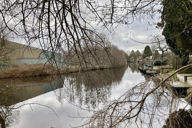Aan de linkerkant van het water de tennishal en de padelbanen, rechts de tuinen van omwonenden.